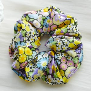 Puede incluir: Un scrunchie negro con estampado floral con flores amarillas, moradas y blancas. El scrunchie está hecho de un tejido suave y elástico.