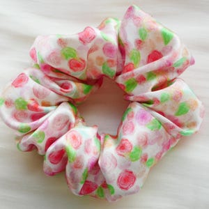 Puede incluir: Un scrunchie blanco con un estampado floral rosa y verde. El scrunchie está hecho de una tela suave y sedosa.