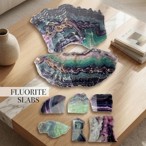 May include: Fluorite slabs in various sizes and shapes, showcasing a spectrum of colors including purple, green, and blue. The slabs are displayed on a wooden surface, with the text "FLUORITE SLABS" visible.