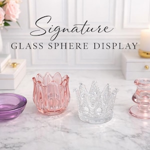 May include: A collection of glass sphere displays, including a purple bowl, a pink tulip-shaped candle holder, a clear crown-shaped holder, and a pink candle holder. The text "Signature GLASS SPHERE DISPLAY" is at the top.