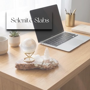 May include: A selenite slab with a clear crystal ball in a gold stand, a selenite tower, and a laptop on a wooden desk. The image also includes a small succulent in a white pot and a ceramic mug.
