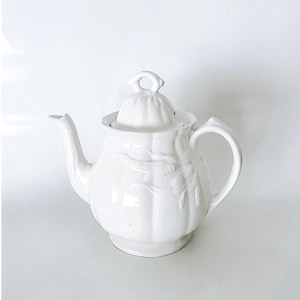 May include: A white ceramic teapot with a lid, spout, and handle. The teapot has a rounded body with embossed floral detailing. The lid has a small loop handle. The teapot is designed for brewing tea.