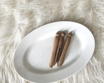 May include: Three wooden clothespins rest on a white oval plate. The clothespins are a warm brown color and have a classic design. The plate is smooth and glossy, and the background is a fluffy, white faux fur.