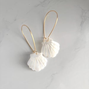 May include: A pair of seashell earrings. Each earring features a white, shell-shaped pendant suspended from a gold-colored, elongated hook. The earrings are displayed on a white and gray marble surface.