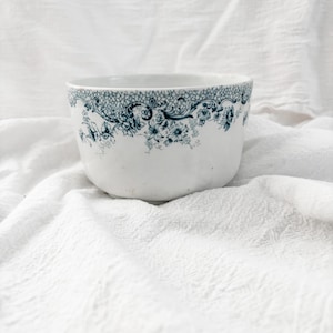 May include: White ceramic bowl with a detailed teal floral and lace pattern around the rim. The bowl has a rounded shape and sits on a white, textured surface. The design is reminiscent of vintage tableware.