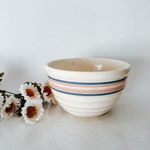 May include: Cream-colored ceramic mixing bowl with horizontal stripes in blue and pink. The bowl has a ribbed exterior and a wide opening. A few white flowers with orange centers are in the background.