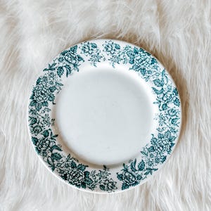 May include: White ceramic plate with a teal floral pattern around the rim. The plate has a simple, elegant design, perfect for serving food. The floral pattern includes detailed leaves and flowers.