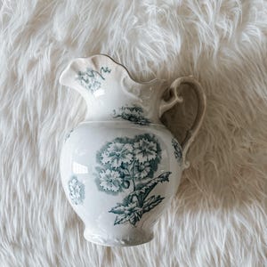May include: White ceramic pitcher with a floral design in teal. The pitcher has a rounded body, a handle, and a ruffled rim. The floral pattern includes large blooms and leaves. A decorative piece for the home.