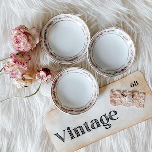 May include: Three small, white vintage dishes with a delicate pink floral border and gold trim. The word "Vintage" is printed on a cream-colored tag, along with the number "68". Pink and cream-colored roses are arranged nearby.