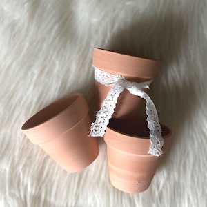 May include: Three small terracotta flower pots with a matte finish. Two of the pots are stacked, and a white lace ribbon is tied around the top of the upper pot, with the ends of the ribbon draped down over the sides of the lower pot.