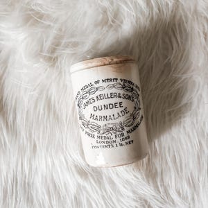 May include: An antique Dundee Marmalade jar with a wooden lid. The white ceramic jar features black text and a decorative emblem. The text reads "JAMES KEILLER & SON'S DUNDEE MARMALADE" and "PRIZE MEDAL FOR LONDON. 1862". The jar contains 1 lb. net.