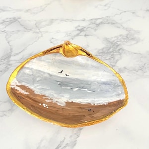 May include: Hand-painted seashell trinket dish. The interior features a beach scene with brown sand, white waves, and a gray sky with two birds. The shell's edge is painted gold.