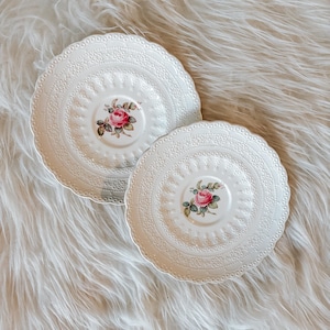May include: Two white ceramic plates with a floral rose design. The plates have a scalloped edge and a detailed embossed pattern. The center of each plate features a pink rose with green leaves.