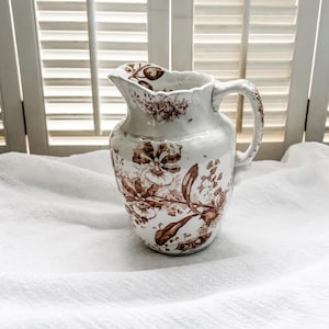May include: A white ceramic pitcher with a brown floral pattern. The pitcher has a wide mouth and a handle.
