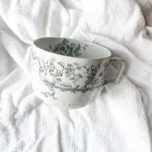 May include: A white ceramic teacup featuring a detailed grey floral pattern that wraps around the exterior and extends into the interior rim. The cup has a curved handle and a smooth, rounded body with a decorative border design.
