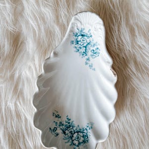 May include: A white ceramic dish shaped like a seashell with scalloped edges and a glossy finish. The dish features delicate teal blue floral patterns printed on the upper and lower sections, with a small brown mark on the right side edge.