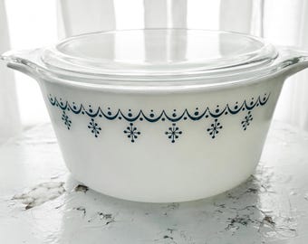 Op de afbeelding: Witte Pyrex-schaal met een doorzichtig glazen deksel. De schaal heeft een blauw sneeuwvlok- en geschulpte rand. De schaal staat op een witte, verweerde kruk.