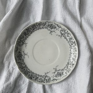 May include: A round ceramic saucer featuring a cream-colored base with a detailed black floral and scrollwork pattern along the scalloped rim. The center of the saucer has a circular indentation designed to hold a cup, with a smooth finish.
