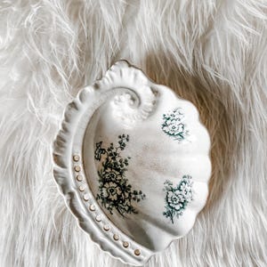 May include: A cream-colored ceramic dish shaped like a seashell, featuring a scalloped edge and a series of small circular indentations along one side. The interior surface is decorated with dark green floral illustrations and a crackled glaze finish.
