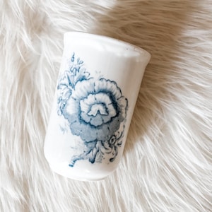 May include: A white ceramic tumbler with a blue floral design. The design features a large, stylized flower with detailed petals and leaves. The tumbler is cylindrical and appears to be vintage or antique.