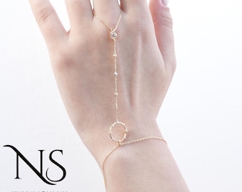Puede incluir: Una delicada cadena de mano dorada con un anillo, que conecta con una pulsera y una cadena que sube hasta el dedo corazón. La cadena está adornada con pequeñas gemas brillantes. El logo "NS KUYUMCULUK" está en la esquina inferior izquierda.