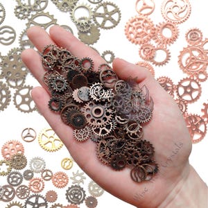 May include: A collection of small, metallic gears in various sizes and shades of bronze, copper, and gold. The gears are held in a hand, with additional gears scattered around. The image has a steampunk aesthetic.