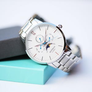 Könnte beinhalten: Eine silberne Armbanduhr mit weißem Zifferblatt und silbernem Armband. Das Zifferblatt hat mehrere Hilfszifferblätter und den Text "architect London". Die Uhr liegt auf einer hellblauen Schachtel.