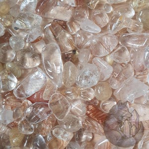 May include: A close-up of a pile of tumbled rutilated quartz crystals. The crystals are a mix of clear and translucent stones with golden and reddish-brown inclusions. The crystals vary in size and shape, creating a natural, organic aesthetic.