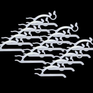 Könnte beinhalten: Eine Ansammlung weißer Kleiderbügel aus Kunststoff mit einem einzigartigen Design. Jeder Bügel hat eine geschwungene Oberseite mit einem dekorativen Element und eine horizontale Stange zum Aufhängen von Kleidungsstücken. Die Bügel sind vor schwarzem Hintergrund angeordnet.