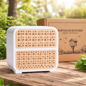 May include: A white and tan bee house with a box in the background. The bee house has two sections with multiple holes. The box has text that reads "SCHÜTZE DIE BESTÄUBER" and "URBANES BIENENHAUS" with a drawing of a bee house.
