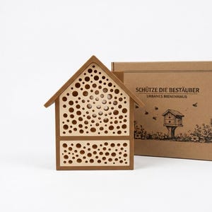 Può includere: Una casetta per insetti in legno con tetto e cornice marroni, caratterizzata da numerose aperture circolari. La casetta è accanto a una scatola di cartone con la scritta "SCHÜTZE DIE BESTÄUBER" e un disegno di una casetta per insetti simile.