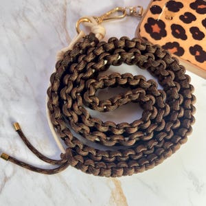 Peut inclure: Une lanière de téléphone en paracorde marron et beige avec un fermoir doré. La lanière est faite de cordon étroitement tissé, créant un design texturé et durable. La lanière est enroulée, montrant sa longueur et sa construction. Un étui de téléphone avec un imprimé léopard est visible.