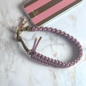 May include: A mauve and olive green phone wrist strap with a gold-colored clasp. The wrist strap is made of woven cord and is attached to a phone case with a pink and brown striped design. The phone case has the text "Coeur Chamallow".