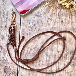 May include: A brown phone lanyard with a gold-colored clasp and a decorative knot. The lanyard features a leopard print accent and is designed to attach to a phone case. The background is a wooden surface.