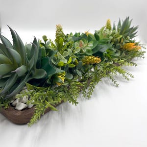 Op de afbeelding: Een decoratieve vetplantenarrangement in een houten trog. Het arrangement bevat verschillende tinten groene vetplanten, kleine witte steentjes en accenten van gele en groene bladeren. De houten trog is donkerbruin van kleur.