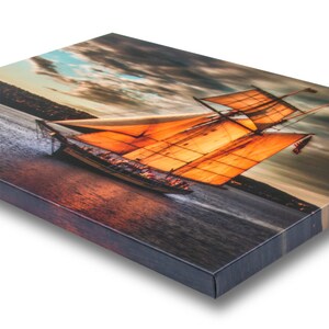 K&ouml;nnte beinhalten: Leinwanddruck mit einem Segelschiff mit orangefarbenen Segeln auf dem Wasser. Das Kunstwerk zeigt einen dramatischen Himmel mit dunklen Wolken und einem Sonnenuntergang. Der Druck ist auf einem Holzrahmen montiert.