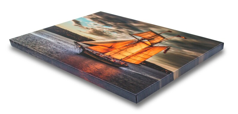 K&ouml;nnte beinhalten: Leinwanddruck mit einem Segelschiff mit orangefarbenen Segeln auf dem Wasser. Das Kunstwerk zeigt einen dramatischen Himmel mit dunklen Wolken und einem Sonnenuntergang. Der Druck ist auf einem Holzrahmen montiert.