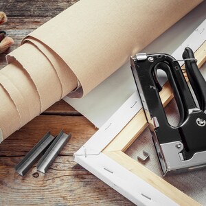 K&ouml;nnte beinhalten: Kunstbedarf auf einer Holzoberfl&auml;che. Abgebildet sind eine Leinwandrolle, eine Tackerpistole, Heftklammern, Pinsel und ein Cuttermesser. Die Leinwand ist beige, die Tackerpistole schwarz und silber.