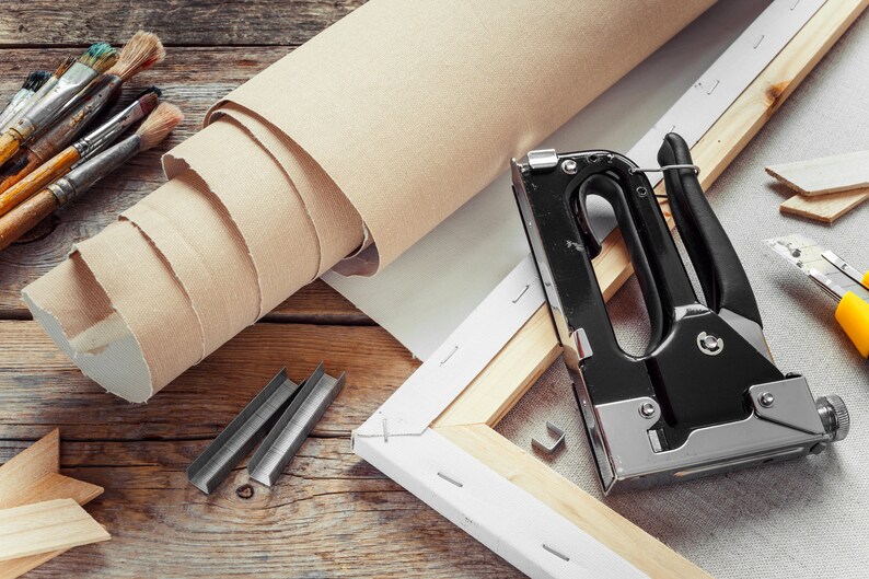 K&ouml;nnte beinhalten: Kunstbedarf auf einer Holzoberfl&auml;che. Abgebildet sind eine Leinwandrolle, eine Tackerpistole, Heftklammern, Pinsel und ein Cuttermesser. Die Leinwand ist beige, die Tackerpistole schwarz und silber.