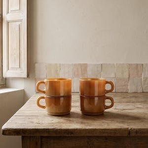 May include: Four amber-colored glass mugs stacked in pairs on a wooden table. Each mug has a rounded body and a curved handle. The mugs are in a warm, inviting color, perfect for serving hot beverages.