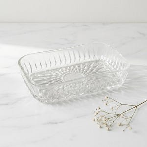 Peut inclure: Un plat rectangulaire en verre transparent avec un motif texturé en forme d'éclat de soleil sur la base. Les côtés présentent des rainures verticales. Une brindille de fleurs blanches est à proximité.
