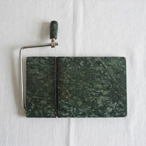 Peut inclure: Une trancheuse à fromage avec une base en marbre vert foncé et un fil et une poignée en acier inoxydable. La poignée est de couleur vert foncé. La trancheuse rectangulaire repose sur une surface blanche.