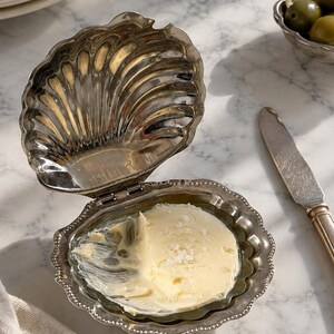 Peut inclure: Un beurrier en métal argenté en forme de coquille Saint-Jacques ouverte avec un couvercle à charnière. L'intérieur de la coquille contient une portion de beurre jaune pâle saupoudrée de cristaux de sel. Les bords présentent une bordure perlée.
