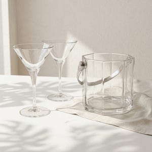 May include: Two clear martini glasses and a clear ice bucket with a metal handle sit on a white surface. The glasses have a classic cone-shaped bowl and a long stem. The ice bucket has a geometric design.
