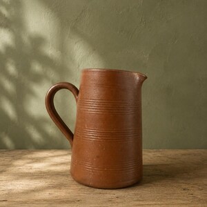 Op de afbeelding: Een terracotta-kleurige keramische kan met een gladde, matte afwerking. De kan heeft een cilindervormig lichaam met drie sets horizontale ingesneden lijnen rondom de buitenkant en een enkele gebogen handgreep aan de zijkant.