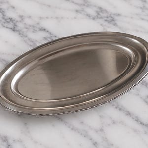 Può includere: Un vassoio da portata ovale in metallo color argento con una base centrale liscia e riflettente e un bordo rialzato con un dettaglio a perline lungo tutto il perimetro esterno. Il vassoio presenta una finitura lucida e un design semplice.