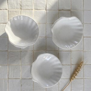 Peut inclure: Trois petits plats blancs en céramique en forme de coquillage sont disposés sur une surface carrelée blanche. Les plats ont un bord festonné et un intérieur lisse. Une tige de blé séchée est placée près de l'un des plats.
