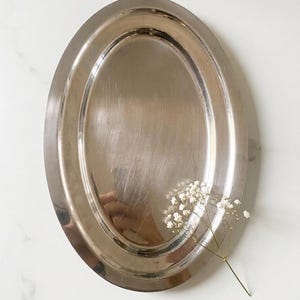 Könnte beinhalten: Ein silberfarbenes, ovales Serviertablett mit poliertem Finish. Das Tablett hat einen erhöhten Rand und eine glatte, reflektierende Oberfläche. Ein Zweig weißer Blumen liegt auf der Oberfläche des Tabletts.