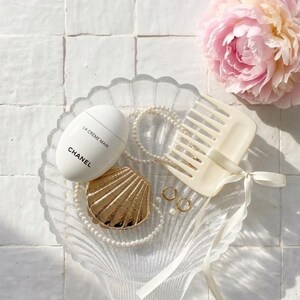 Peut inclure: Un plat transparent en forme de coquillage contient une crème pour les mains Chanel blanche, un poudrier doré en forme de coquillage, un collier de perles, des créoles dorées et un peigne crème avec un ruban. Une pivoine rose est également présente.