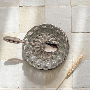Peut inclure: Une cuillère en argent repose dans une soucoupe en verre décorative avec un motif géométrique texturé. Le plat est posé sur une surface carrelée blanche, avec un seul brin de blé sur le côté. La cuillère et le plat sont réfléchissants.